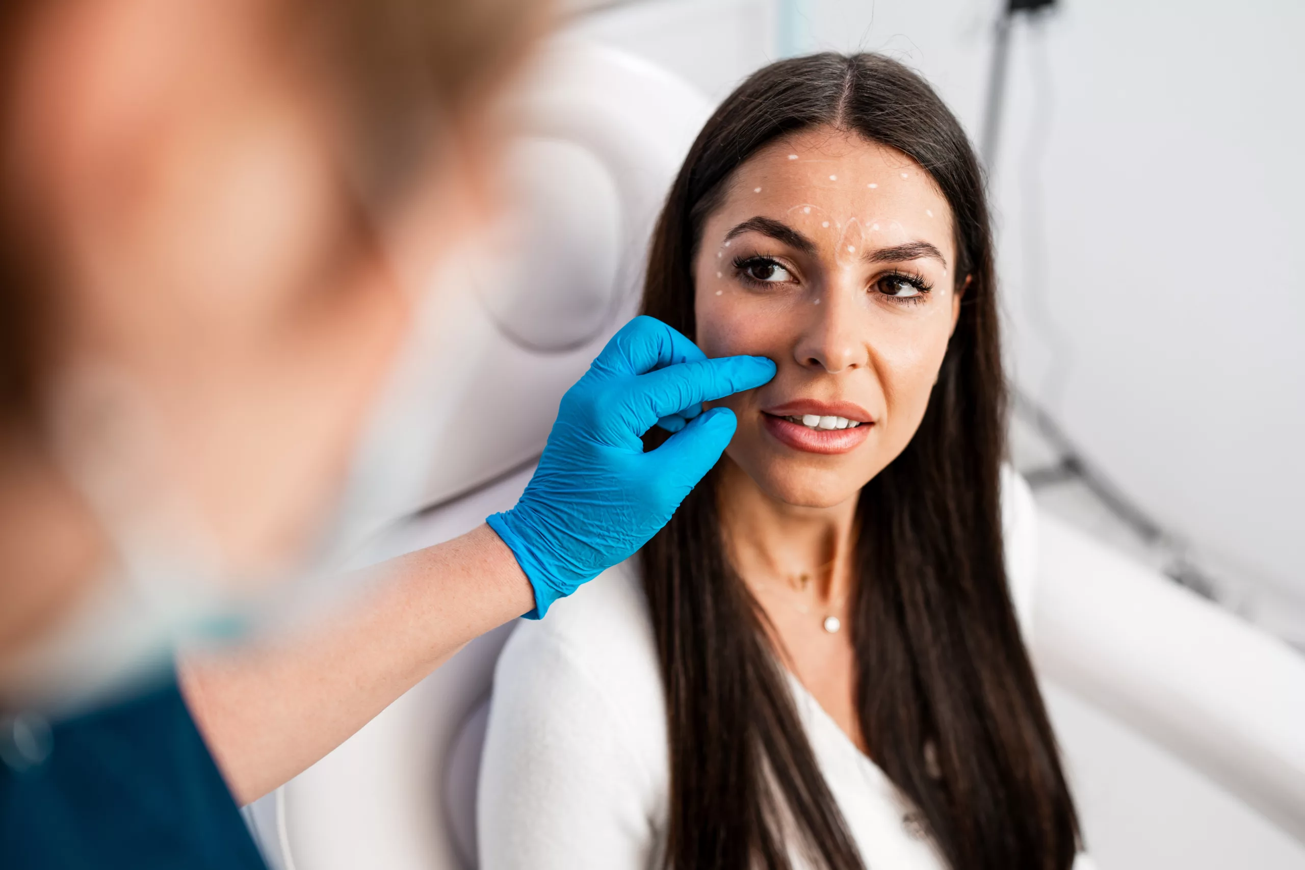 Beautiful and happy brunette woman at beauty medical clinic. She is sitting and talking with female doctor about face aesthetic treatment.